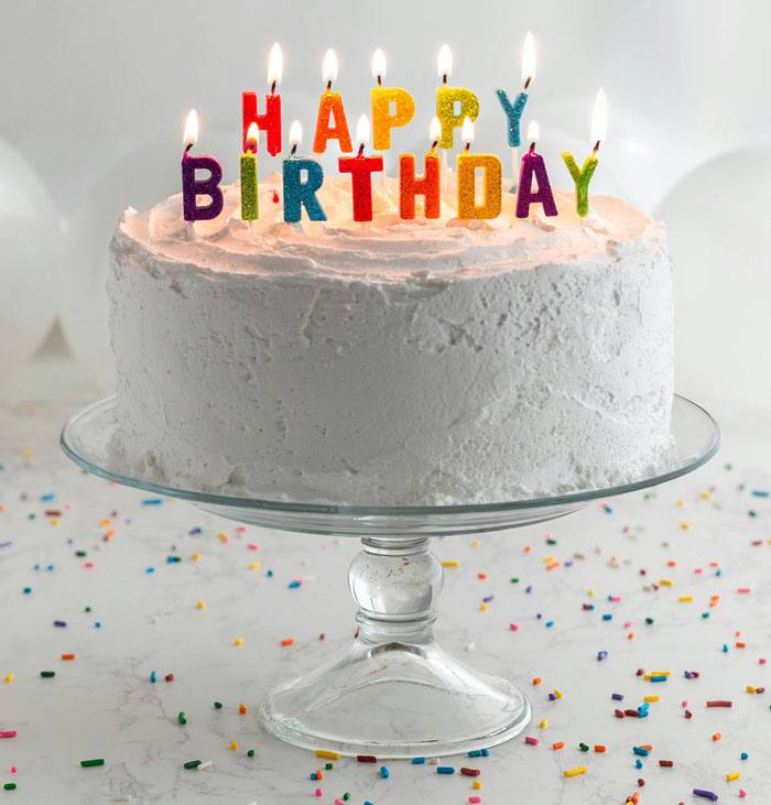 Birthday cake with colorful lit candles spelling happy birthday on a glass stand with confetti sprinkles nearby