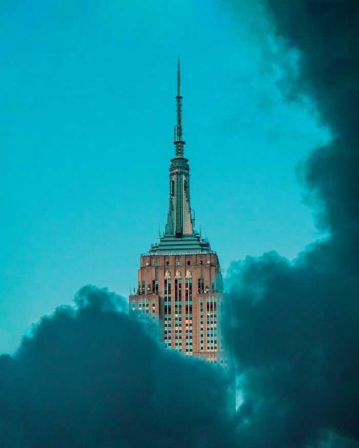 Surreal photo manipulation of a skyscraper emerging through thick clouds under a vivid turquoise sky.