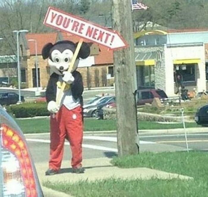 Person in a creepy Mickey Mouse costume holding a sign that says You’re Next in a suburban street, cursed image.