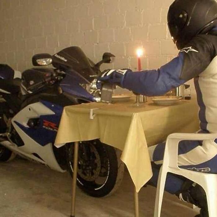 Motorcyclist wearing helmet and gloves, having dinner at a table set next to a parked motorcycle, a surreal cursed image.