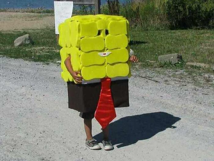Child wearing an oversized yellow box costume with a red tie standing on a gravel road, a cursed image for weird breaks.