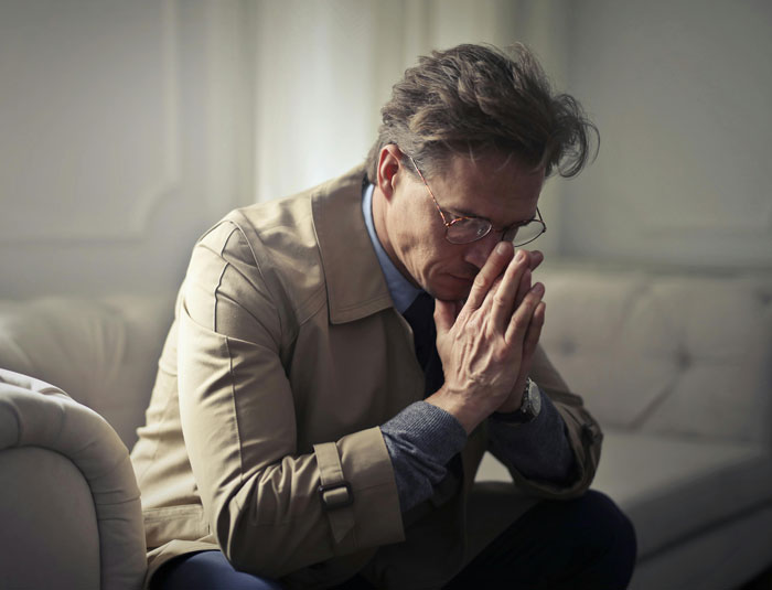 Man in glasses and trench coat sitting on a couch looking stressed, reflecting on infuriating things said by people in charge.