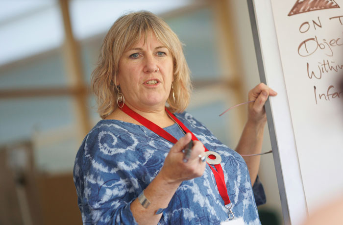Woman in blue tie-dye shirt holding glasses and pointing to a whiteboard with infuriating things said by people in charge.