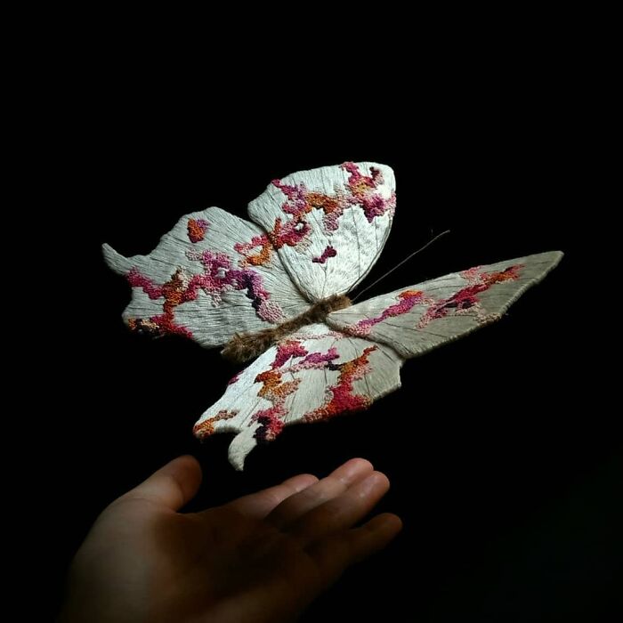 Hand holding a detailed embroidered fiber moth with pink and orange stitches on white wings against black background.