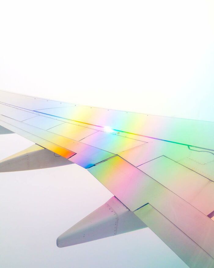 Airplane wing with creative photo manipulations showing a surreal rainbow light effect in a bright sky view.