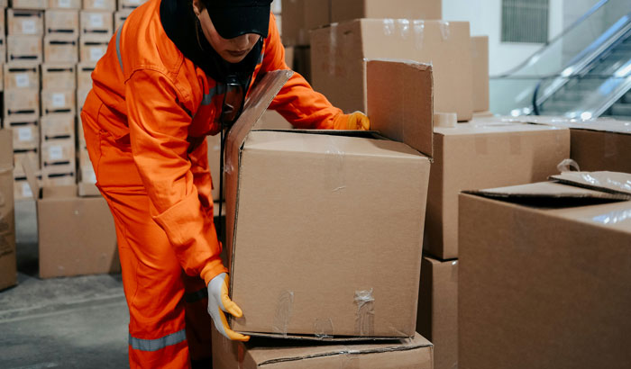 Warehouse worker in orange uniform handling large cardboard boxes near empty shelves inside storage facility Warehouse worker in orange uniform handling large cardboard boxes near empty shelves inside storage facility