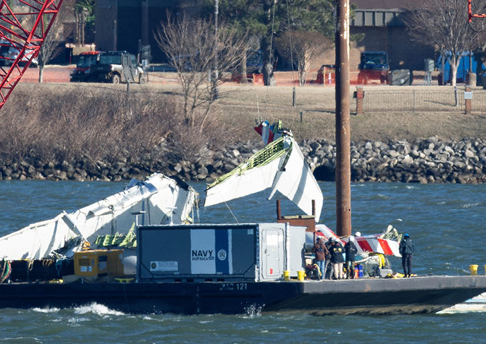 Wreckage of a DC plane crash on water with Navy salvage crew examining the debris near a shore with vehicles and trees. Wreckage of a DC plane crash on water with Navy salvage crew examining the debris near a shore with vehicles and trees.