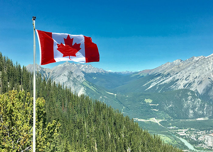 Canadian flag waving on a pole with a backdrop of mountains and forest, reflecting life in home countries experiences.