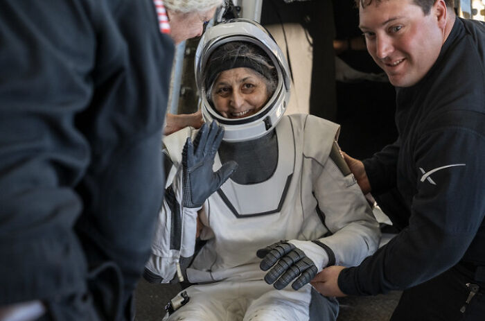Astronaut in space suit being assisted by two people, showing struggles faced after long time in space.
