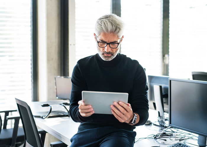 Middle-aged man in glasses using tablet in modern office, reflecting on infuriating things said by people in charge.