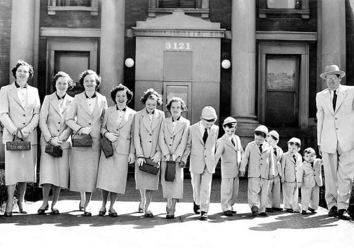 Group of women and children dressed in vintage clothing standing in front of a historic building in an antique historical photograph.