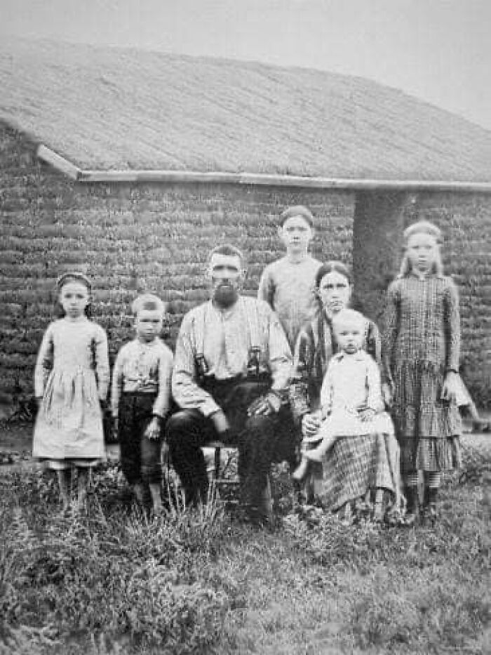 Black and white antique historical photograph of a family with five children in front of a rustic building.