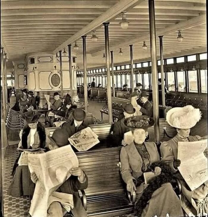 Antique historical photograph of early 20th-century passengers reading newspapers on a vintage train carriage.