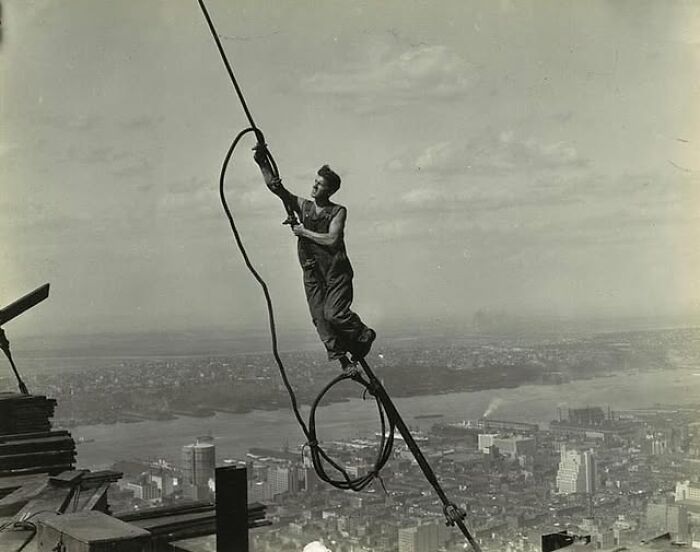 Man balancing on a cable high above a city during construction in an antique historical photograph.