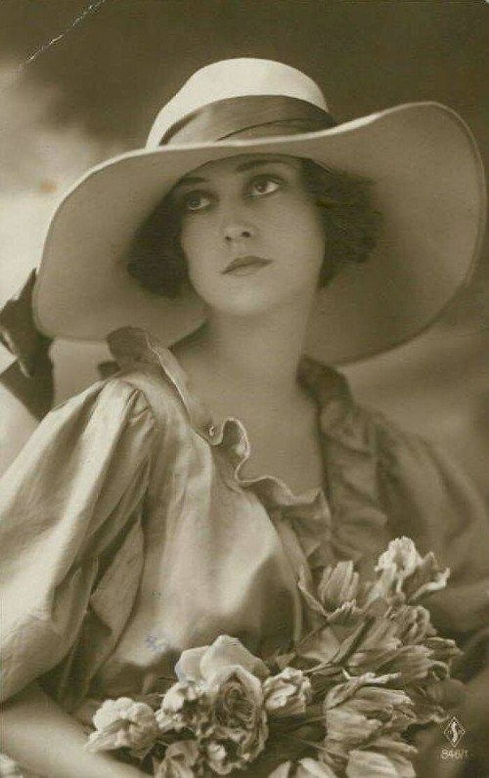 Vintage antique historical photograph of a woman in a wide-brimmed hat holding a bouquet of flowers in sepia tone.