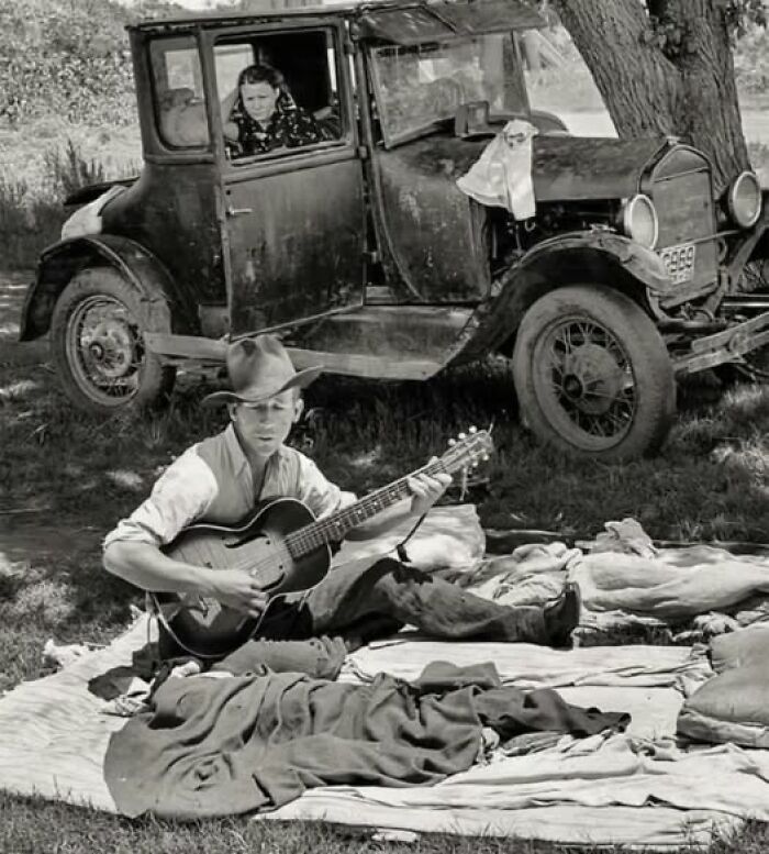 Man playing guitar outdoors by an old car in an antique historical photograph showing life in the past.