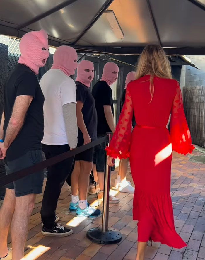 Woman in red dress walking past men in pink ski masks, illustrating bleeding and hospital update for adult content creator. Woman in red dress walking past men in pink ski masks, illustrating bleeding and hospital update for adult content creator.