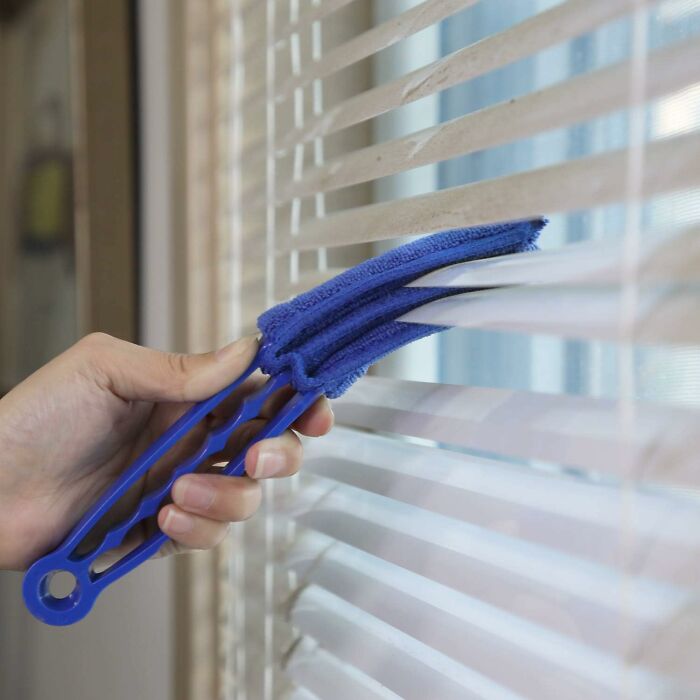 Hand using a blue cleaning tool to dust window blinds, illustrating practical finds for people who want to nail adulting.
