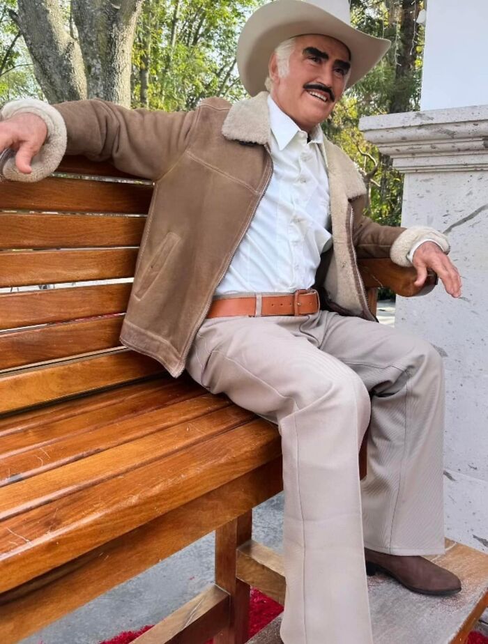 Hyper-realistic sculpture by Ruben Orozco Loza seated on a wooden bench wearing a cowboy hat and brown jacket.
