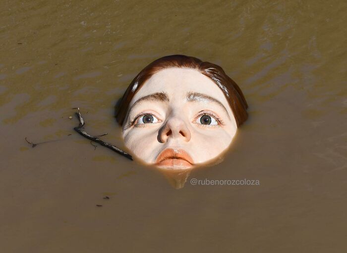 Hyper-realistic sculpture by Ruben Orozco Loza of a lifelike female face partially submerged in muddy water.