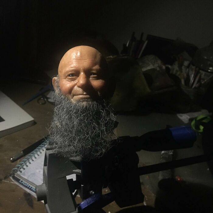Hyper-realistic sculpture head with detailed beard and lifelike facial features by Ruben Orozco Loza in dim workshop lighting.