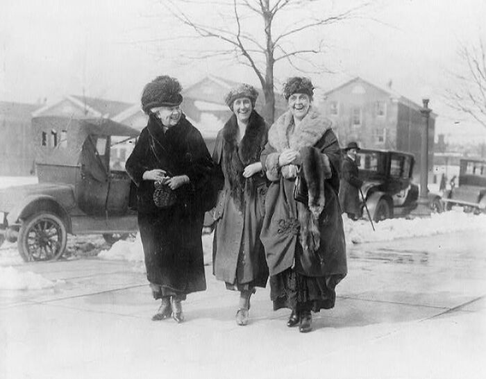 Three women dressed in early 1900s fashion walking on a snowy American street, showcasing life in America from 100 years ago.