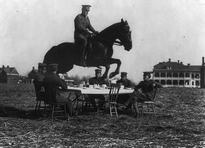 Men in vintage military uniforms sit at a table outdoors while a man on horseback jumps over them in life in America 100 years ago.