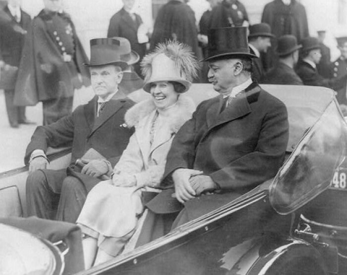 Three people dressed in early 20th century attire riding in a vintage car showcasing life in America 100 years ago.