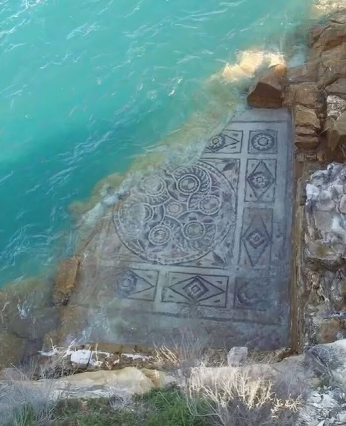 Ancient mosaic partially submerged in turquoise water, showcasing intricate patterns near rocky shoreline from archaeology news.