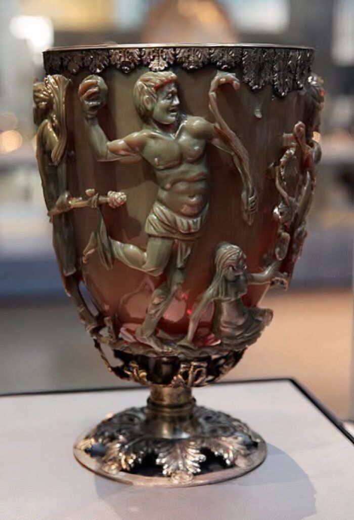 Ancient ornate cup with detailed carved figures on bronze surface displayed in an archaeology museum setting.