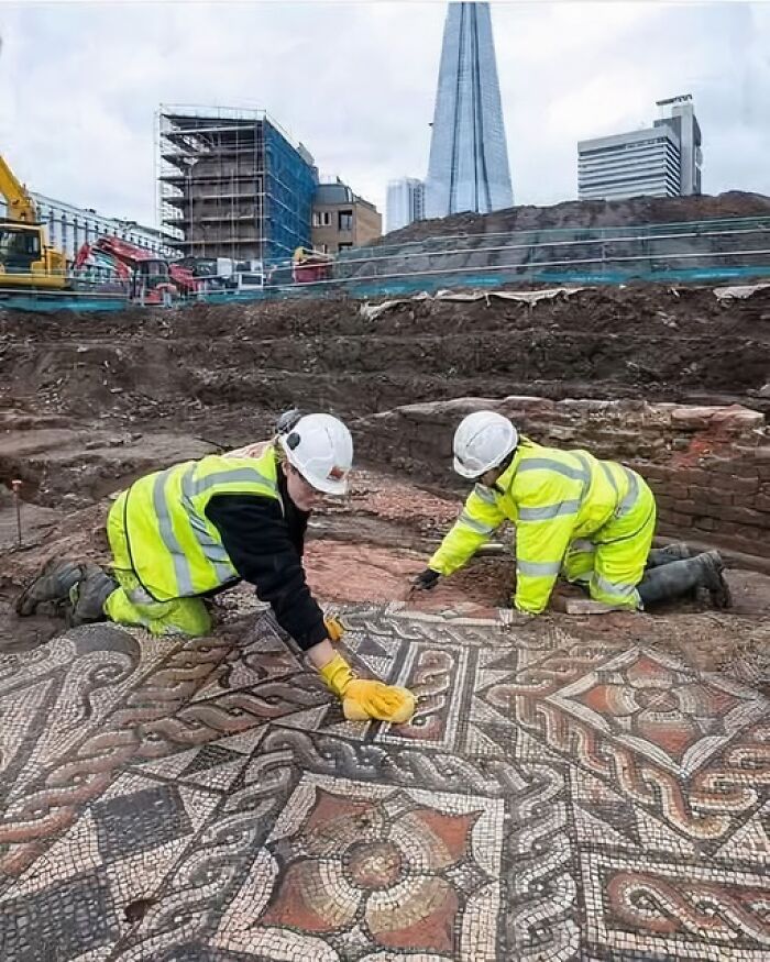 Archaeologists cleaning an intricate mosaic floor at an urban excavation site with modern buildings in the background.