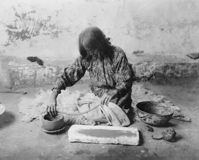Native American woman in the 1900s preparing traditional crafts inside a rustic room with handmade tools around her.