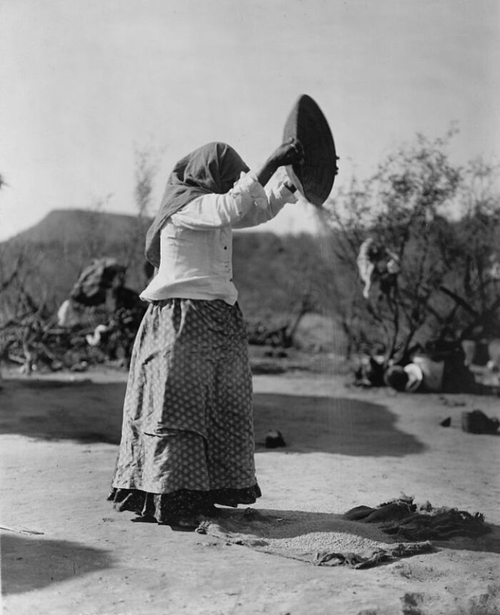 Native American woman in traditional dress sifting grain outdoors in a rare and powerful photo from the 1900s.