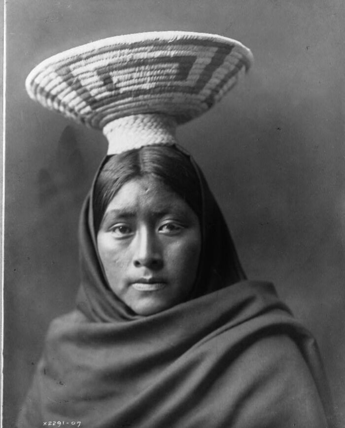 Native American woman from the 1900s wearing traditional woven basket on her head and wrapped in a blanket.