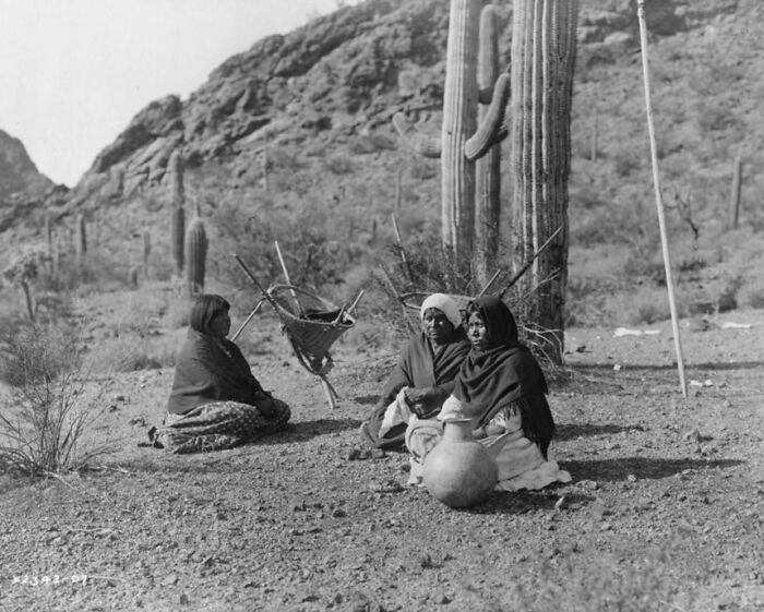 Three Native American women sitting on the ground near cacti in the desert, depicting life in the 1900s.