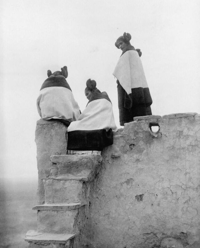 Three Native American women wrapped in blankets sitting on adobe steps, revealing Native American life in the 1900s.