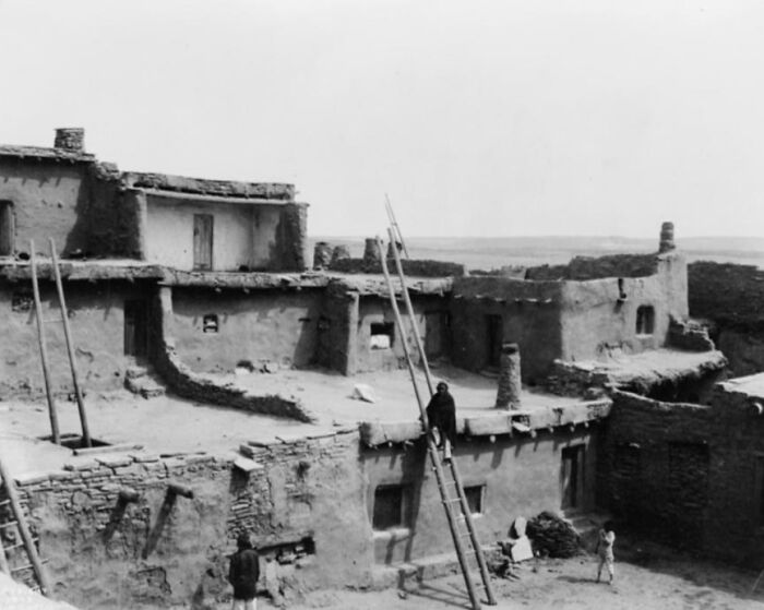 Black and white photo showing Native American adobe homes and people using ladders in early 1900s Native American life.
