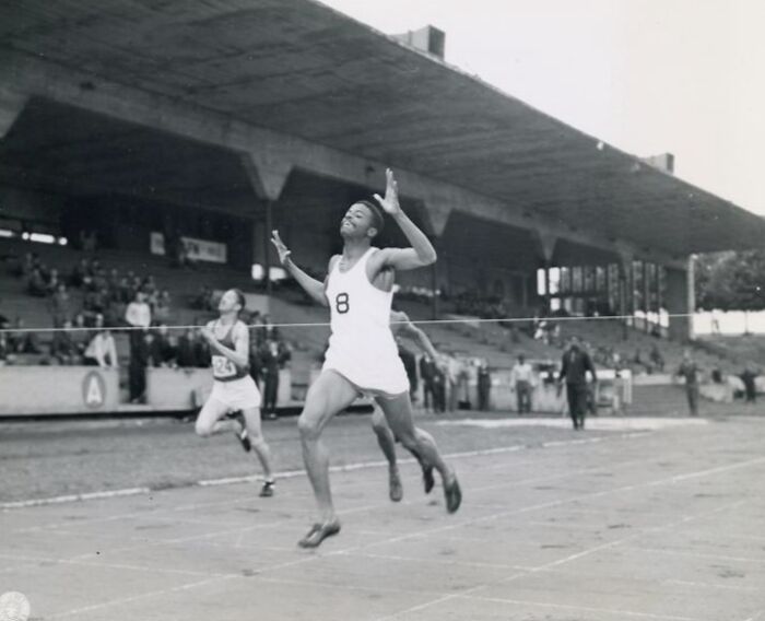 African American athlete in uniform crossing finish line during historical track race at outdoor stadium with spectators.