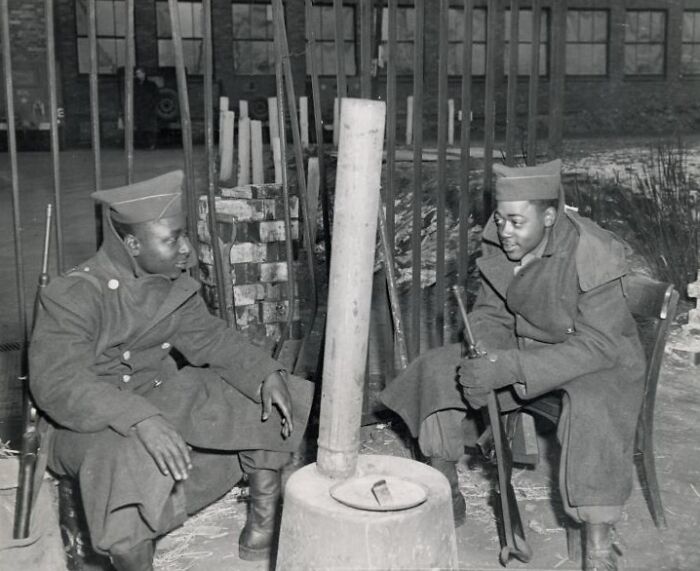 Two African American soldiers in uniform sitting and warming by a stove, historical black and white photo.