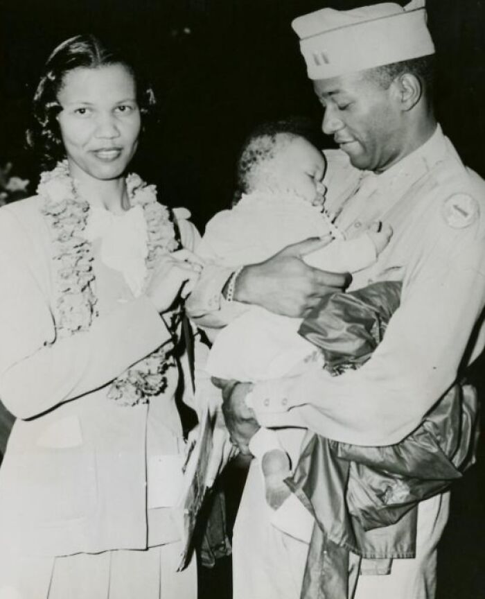 African American man in military uniform holding baby while woman stands beside them wearing a floral lei.