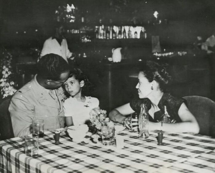 African American man in military uniform sitting at a table with a woman and child in a vintage black and white photo.