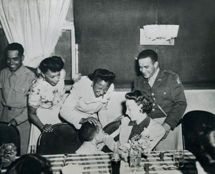 African Americans in uniform share a joyful moment with family around a dining table in a historic black and white photo.