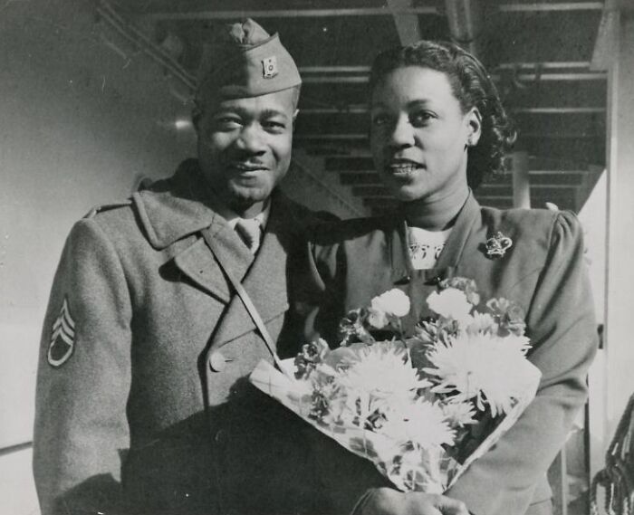 African American soldier in uniform standing next to a woman holding a bouquet of flowers in a historic black and white photo.