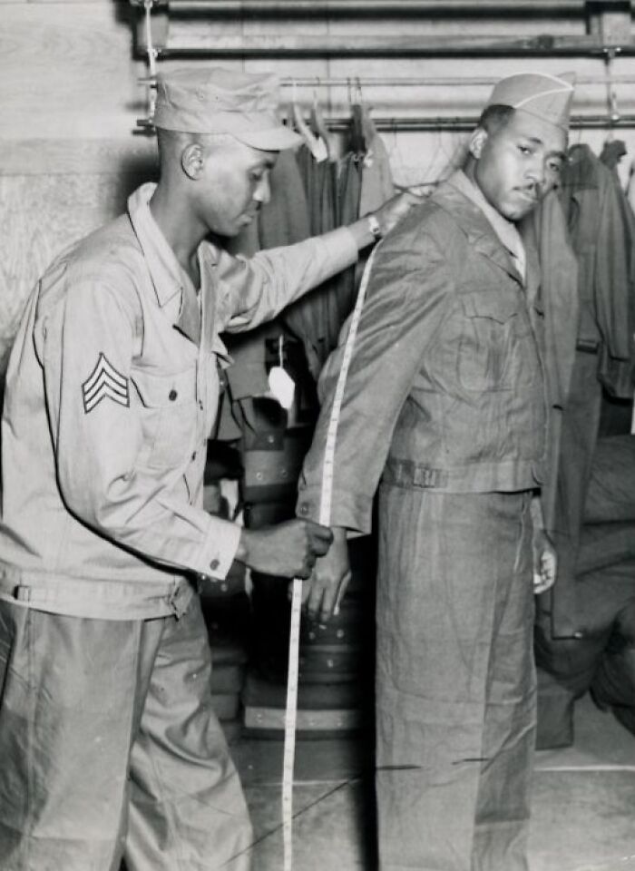Two African American soldiers in uniform, one measuring the other's sleeve length, showcasing historical military attire.