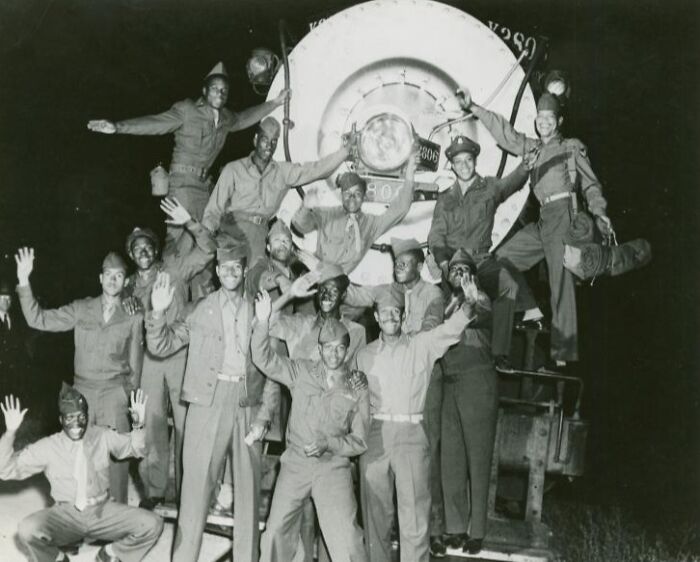 Group of African Americans in uniform posing happily in front of a large train engine at night in a historic black and white photo
