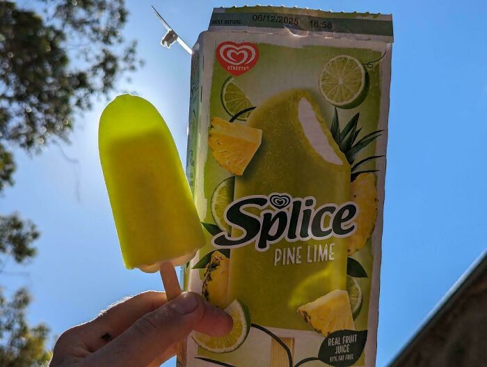 Hand holding a small Splice Pine Lime frozen treat next to its larger packaging under blue sky showing product size difference.