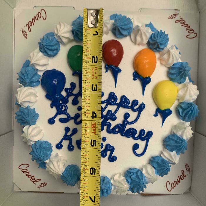 Small birthday cake with colorful balloons and blue and white icing, measured with a tape showing its size.