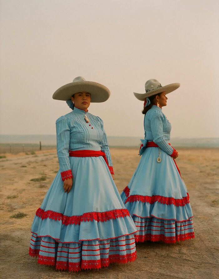 Two all-female rodeo riders in traditional blue and red dresses and wide-brimmed hats standing in a desert landscape.