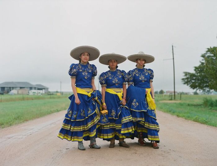 Three all-female rodeo riders in traditional blue dresses and wide-brimmed hats standing on a rural road.