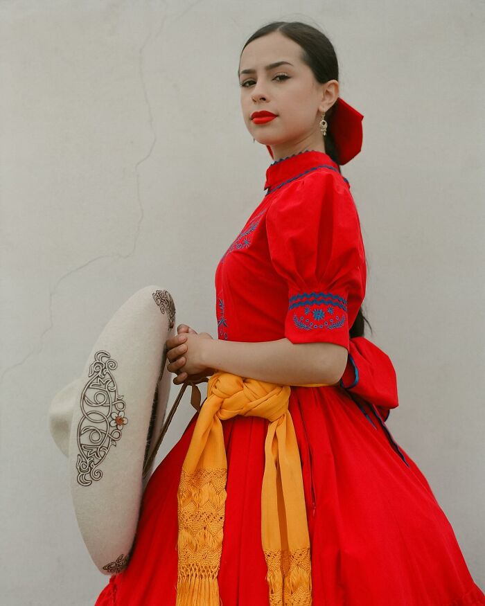 Female rodeo rider in vibrant red traditional dress holding embroidered hat, showcasing the beauty and power of all-female rodeo riders.